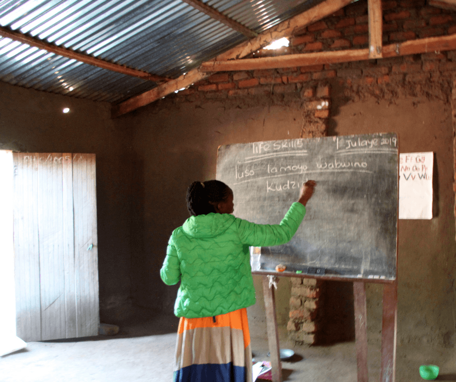 A teacher, wearing a green jacket, writes on a chalkboard in a classroom. Her back is to the camera. 
