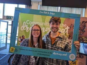 Two Flourish 2023 festival attendees hold a frame which reads 'I'm not a hero. I believe it's time to put communities in the driving seat of development'