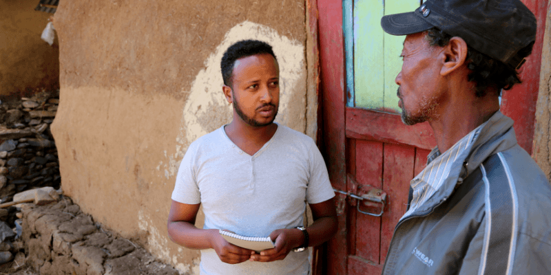 One of All We Can's local partner staff talks to a Village Chairman in Ethiopia, standing outside a homestead with a red and teal front door.