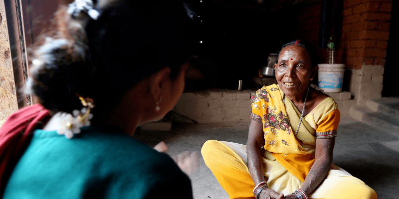 Manato, dressed in a yellow Sari, sits cross legged opposite an employee of one of All We Can's local partners in India