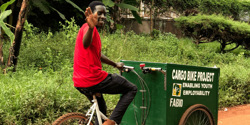 Bernard, in a red top and dark trousers, waves as he cycles on an adapted cargo bike.