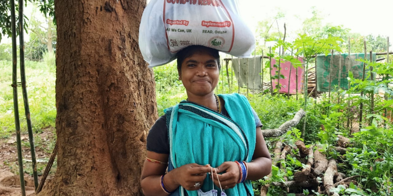 A woman in a blue sari smiles at the camera. Emergency food rations and sanitary equipment are in a bag balanced on her head. In the background is a tree and some washing.