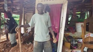 Noel, in a blue top and trousers, stands in the ruin of his home in Haiti. 