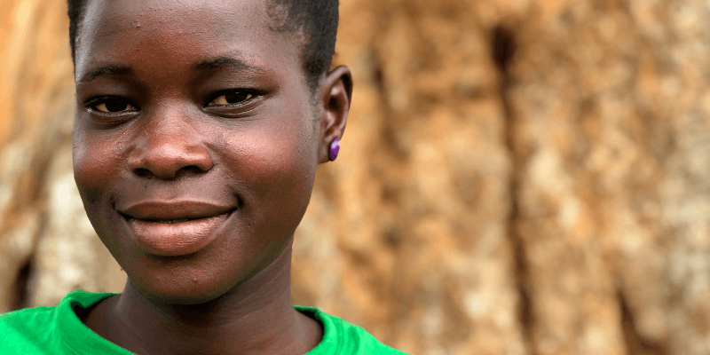 A young woman in a bright green t-shirt stands in front of a tree. She smiles directly into the camera.